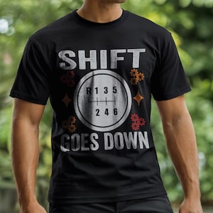 Puede incluir: Camiseta negra con la frase "SHIFT GOES DOWN" en fuente blanca desgastada. El diseño incluye un patrón de cambio de marchas con los números 1, 2, 3, 4, 5 y 6. Gráficos de engranajes y diamantes naranjas y rojos rodean el patrón.