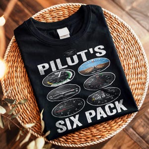 Può includere: T-shirt nera con la scritta bianca "PILOT'S SIX PACK", con illustrazioni di indicatori di strumenti aeronautici. La maglietta è esposta su una superficie circolare intrecciata, creando un look casual.