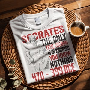 May include: White t-shirt featuring a graphic of Socrates and the quote "The only true wisdom is in knowing you know nothing." The text is in red and black. A cup of coffee sits to the right of the shirt.