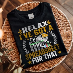 Può includere: T-shirt nero con la scritta "RELAX I'VE GOT A SPREADSHEET FOR THAT" in bianco e giallo. La grafica include un grafico a torta, un grafico a linee e una griglia di foglio di calcolo. La maglietta è piegata su un cestino intrecciato.