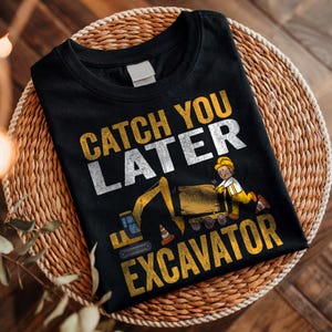 May include: Black t-shirt with the text "CATCH YOU LATER EXCAVATOR" in yellow and white. The design includes a cartoon excavator and a construction worker. The shirt is displayed on a woven, circular placemat.