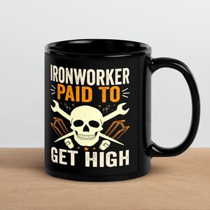 Op de afbeelding: Zwarte keramische mok met een witte schedel en gekruiste moersleutels. De mok heeft de tekst "IRONWORKER PAID TO GET HIGH" in wit en oranje. De mok staat op een lichtbruin oppervlak.
