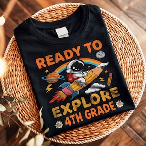 May include: A black t-shirt featuring a graphic of an astronaut riding a yellow pencil like a rocket. The text reads READY TO EXPLORE 4TH GRADE. The design includes a planet with rings, a rainbow, a moon, a small pencil, and two white flowers.