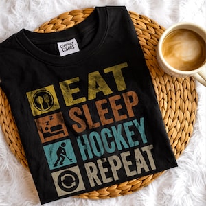 May include: Black t-shirt with the words "EAT SLEEP HOCKEY REPEAT" in a retro font. The design includes icons of a fork and spoon, a bed, a hockey player, and a recycling symbol. A coffee mug sits to the right.
