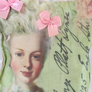 Puede incluir: Una pequeña almohada decorativa con una imagen vintage de una mujer con un lazo rosa en el pelo. La almohada está adornada con cuentas rosas y tiene un fondo verde con texto escrito a mano. El texto dice "Jenge Claustryme 1735 en la nuit".