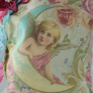 May include: A cream-coloured pillow featuring a vintage illustration of an angel holding a crescent moon. The pillow is adorned with pink and blue ribbons and a beaded fringe. The text "Farfalla Desi" is printed on the pillow.