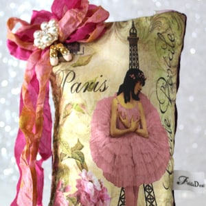 Puede incluir: Una almohada rosa y blanca con un tema parisino vintage. La almohada presenta una bailarina con un tutú rosa, la Torre Eiffel y las palabras "París" y "Carte Postale". La almohada tiene una cinta rosa con un adorno de perlas y una franja de cuentas.