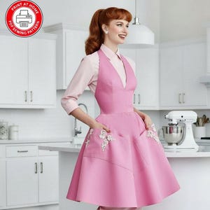 May include: A pink apron with a V-neckline, a flared skirt, and two large front pockets featuring white floral lace appliqués. The apron is worn over a light pink collared shirt and a matching skirt. A red circular icon with a printer and text reads PRINT AT HOME PDF SEWING PATTERN.