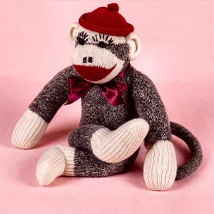 May include: A stuffed monkey toy crafted from dark gray and cream-colored knit fabric. The monkey features a red knit cap with a small pom-pom, a dark red satin bow at the neck, and embroidered facial details including eyes and a mouth.