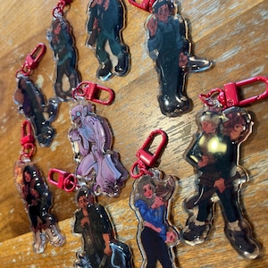 Puede incluir: Una colecci&oacute;n de llaveros acr&iacute;licos transparentes con ilustraciones de personajes. Cada llavero tiene un mosquet&oacute;n rojo para sujetar. Los personajes se representan en varias poses y atuendos, con una gama de colores y estilos.