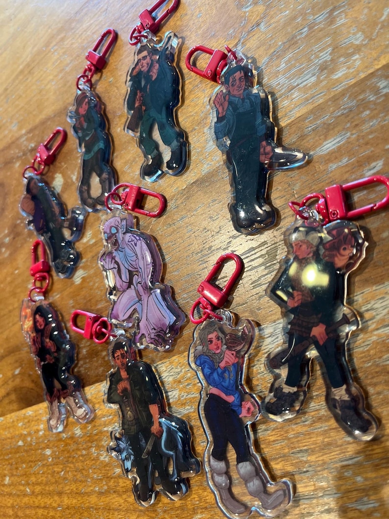 Puede incluir: Una colecci&oacute;n de llaveros acr&iacute;licos transparentes con ilustraciones de personajes. Cada llavero tiene un mosquet&oacute;n rojo para sujetar. Los personajes se representan en varias poses y atuendos, con una gama de colores y estilos.