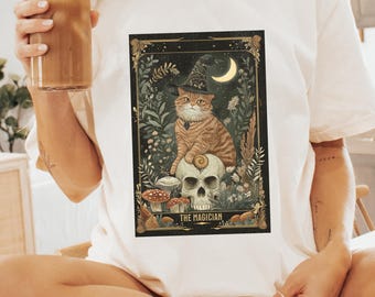 May include: Ivory t-shirt with a detailed illustration of a cat in a witch hat, sitting on a skull, with the text "THE MAGICIAN." The design includes a crescent moon and botanical elements. The shirt is a mock-up for advertising purposes.