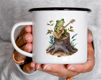 May include: White enamel mug with a black rim featuring a green frog playing a guitar on a tree stump. The frog is sitting on the stump with its legs crossed and holding a guitar in its hands. The guitar is brown with a light brown pickguard. The frog is surrounded by green leaves and brown rocks.
