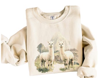 Puede incluir: Una sudadera de color crema con un diseño de acuarela de dos alpacas blancas frente a una cordillera. Las alpacas están de pie en un área cubierta de hierba con una estructura de piedra detrás de ellas.