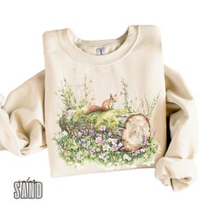 May include: A beige sweatshirt featuring a printed design of a squirrel perched on a tree trunk, surrounded by a variety of pink, purple and white flowers.