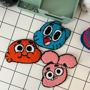 May include: Three colorful felt characters from the animated series The Amazing World of Gumball. The characters are Gumball, Darwin, and Anais. Gumball is blue, Darwin is orange, and Anais is pink.