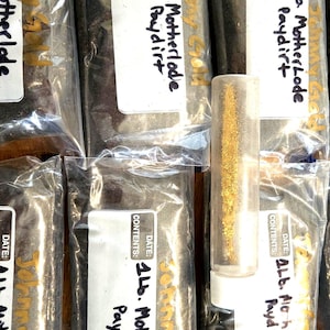 May include: Four clear plastic bags filled with dark brown dirt labeled "1 lb. Motherlode Paydirt" and "Johnny Gold". A small clear glass vial filled with gold flakes is in the center of the image.