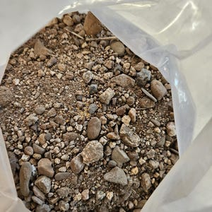 May include: A close-up of a plastic bag filled with a mixture of small and large rocks, gravel, and dirt. The rocks are various shades of brown, gray, and white.