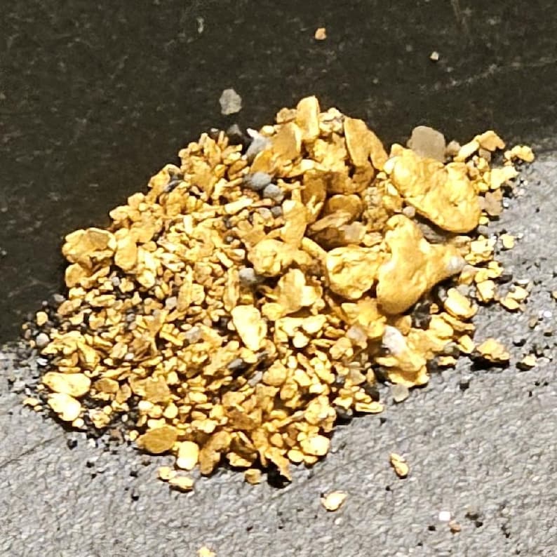 May include: A close-up of a pile of gold nuggets and flakes. The gold is a bright yellow color and is scattered on a dark gray surface.