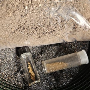 May include: Two small glass vials containing gold nuggets and gold dust. The vials are sitting on a black surface with a green circular pattern.