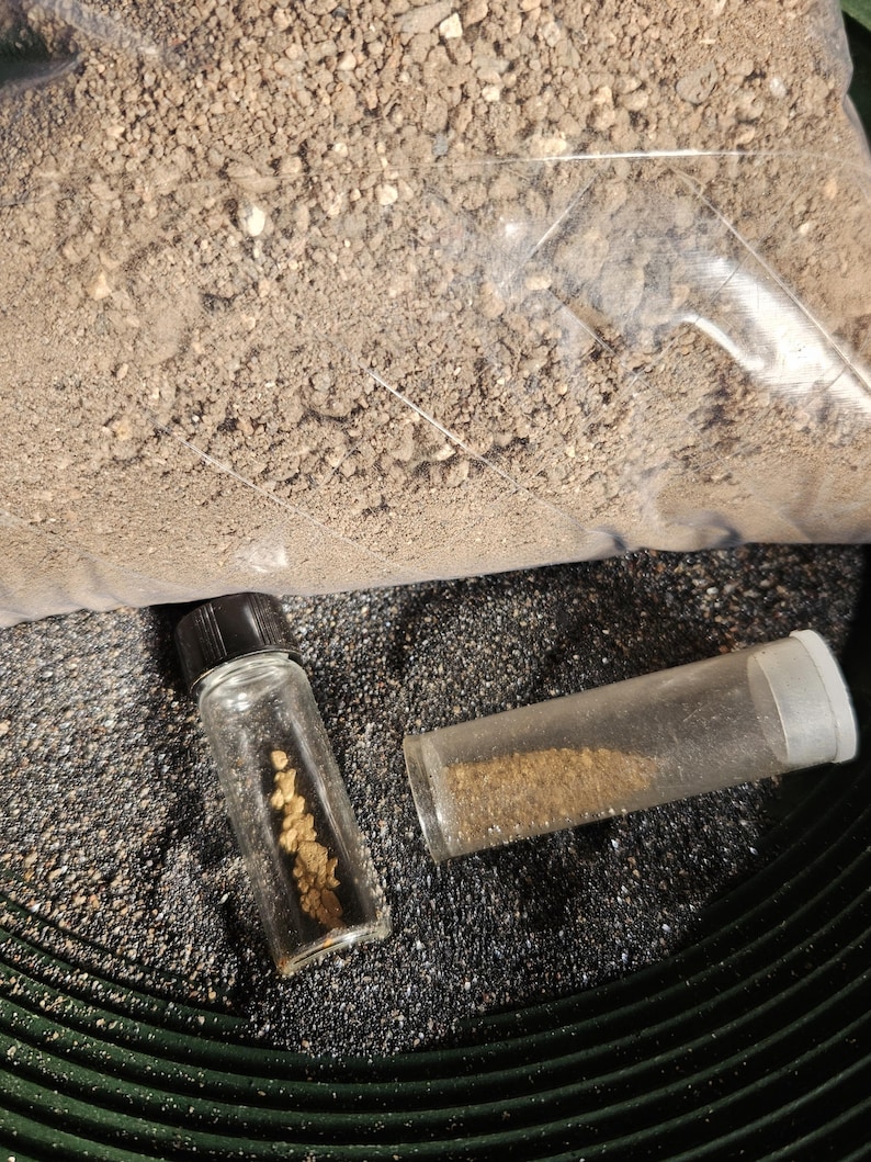 May include: Two small glass vials containing gold nuggets and gold dust. The vials are sitting on a black surface with a green circular pattern.