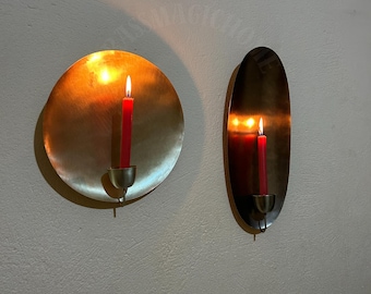 Op de afbeelding: Twee messing wandkandelaars, een ronde en een ovale, elk met een rode kaars met een vlam.