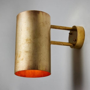 May include: A cylindrical wall sconce with a gold finish. The sconce has a round base and two horizontal arms connecting to a cylindrical shade. The interior of the shade has an orange glow. The fixture is mounted on a white wall.
