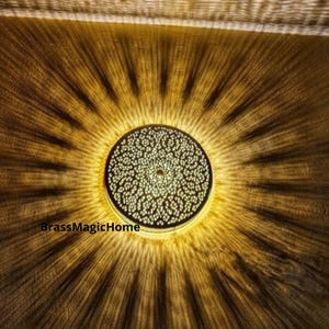 May include: A round, brass ceiling light fixture with a decorative, intricate pattern. The light is on and casting a warm glow.