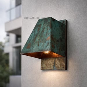 May include: A wall-mounted outdoor light fixture with a weathered, turquoise-green finish. The fixture has a rectangular base and a sloped, square shade. The interior of the shade is a warm, copper color, and the light bulb is visible. The text "BRASSMAGICHOME" is visible.