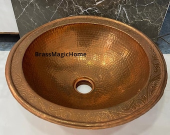 May include: A round copper sink with a hammered finish and an ornate design around the edge. The sink has a drain hole in the center.
