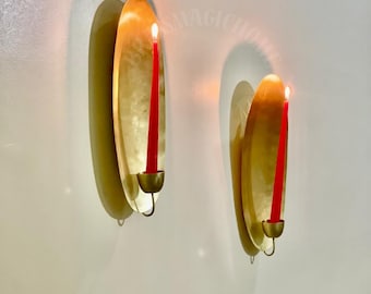 Op de afbeelding: Twee ovaalvormige wandkandelaars van goudkleurig metaal, elk met een glad, reflecterend oppervlak en een kleine komvormige houder. In elke houder staat een slanke rode dinerkaars met een brandende vlam.