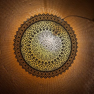 Pode incluir: Uma luminária de teto redonda de metal preto com um design intrincado e rendado. A luz está acesa e projeta um brilho quente.