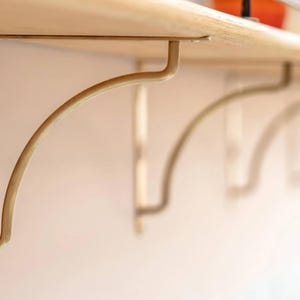 Könnte beinhalten: Nahaufnahme eines Holzregals, das von gebogenen, goldfarbenen Metallhalterungen getragen wird. Die Halterungen sind an einer hellen Wand befestigt und schaffen ein minimalistisches und funktionales Design. Das Regal scheint aus hellem Holz zu bestehen.