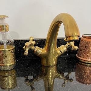 May include: A gold-toned brass faucet with two handles. The faucet has a traditional design with a curved spout and cross-shaped handles. The faucet is mounted on a black countertop.