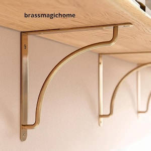 Può includere: Staffe dorate per mensole con un design curvo, montate su una parete di colore chiaro. Le staffe sostengono una mensola in legno. Il testo "brassmagichome" è visibile sulla mensola.