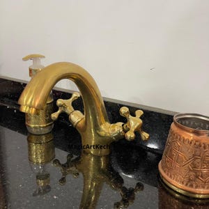 May include: A gold-colored bathroom faucet with cross-shaped handles. A matching soap dispenser and a copper-colored cup with an ornate design are also visible. The faucet has a curved spout and is set against a black countertop.
