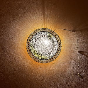 Op de afbeelding: Een ronde, decoratieve plafondlamp met een ingewikkeld, kant-achtig ontwerp. Het licht is aan en geeft een warme gloed.