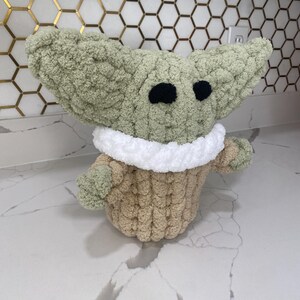 May include: A green and brown crocheted plush toy shaped like a baby alien with large ears and big eyes. The toy is sitting on a white countertop.