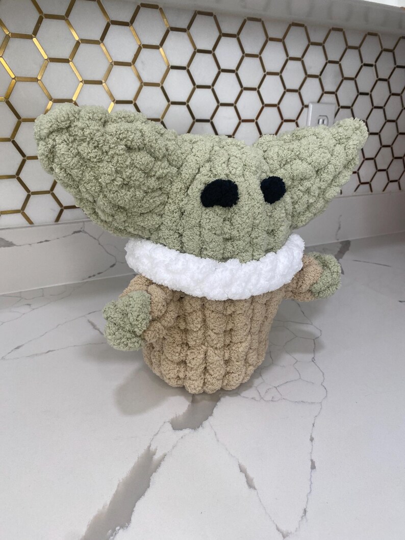May include: A green and brown crocheted plush toy shaped like a baby alien with large ears and big eyes. The toy is sitting on a white countertop.