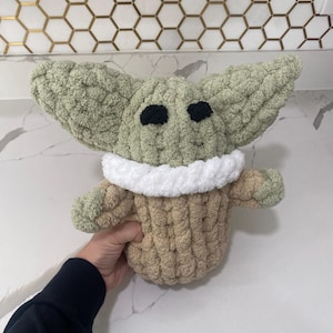 May include: A crocheted plush toy in the shape of a green alien character wearing a brown and white sweater. The toy has large black eyes and a small mouth.