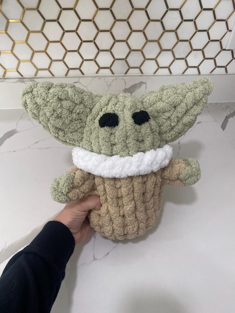 May include: A crocheted plush toy in the shape of a green alien character wearing a brown and white sweater. The toy has large black eyes and a small mouth.