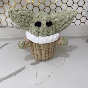 May include: A green crocheted plush toy with large ears, black eyes, and a white and tan crocheted collar. The toy is sitting on a white countertop.
