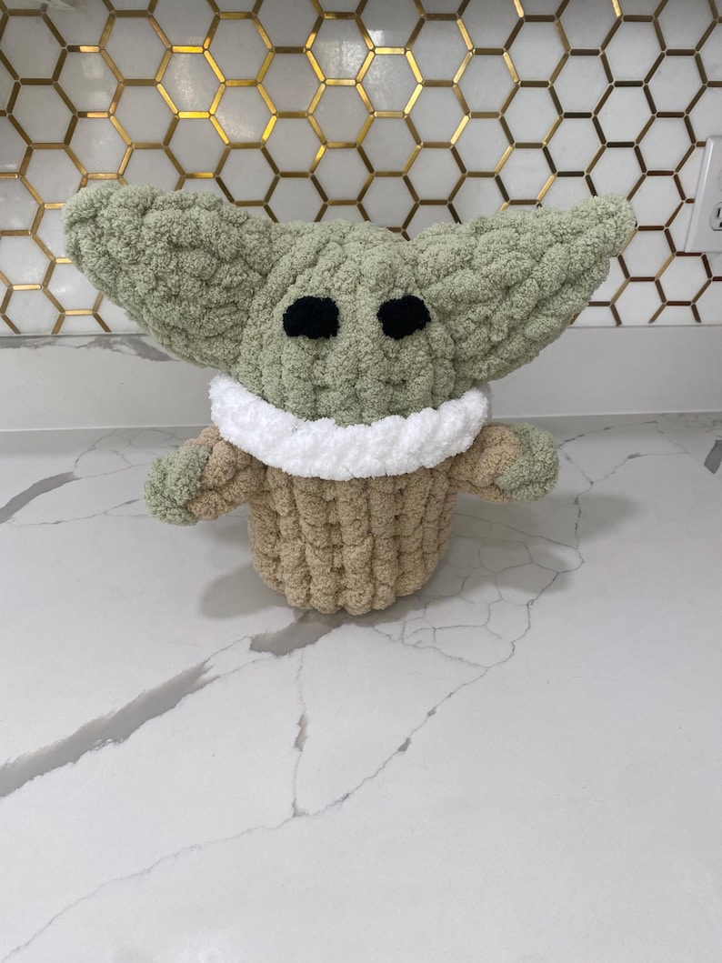 May include: A green crocheted plush toy with large ears, black eyes, and a white and tan crocheted collar. The toy is sitting on a white countertop.