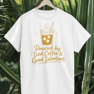 Pode incluir: Uma t-shirt branca de manga curta com um gráfico dourado de um copo de café gelado com palhinha e cubos de gelo. Abaixo do gráfico, o texto "Powered by Iced Coffee & Good Intentions" está escrito numa combinação de letra cursiva e de imprensa.