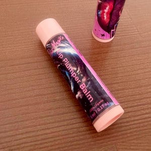 May include: Pink Magik Lip Plumper Balm tube with a pink cap and a label featuring a cosmic design. The label also includes the text "100% Organic" and "5 ml / 0.17 oz". Another tube is visible in the background.