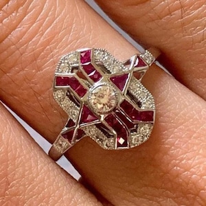 Peut inclure: Une bague en argent avec un grand diamant central entouré d'un motif géométrique de rubis rouges et de petits diamants.