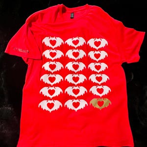 May include: Red t-shirt with a repeating pattern of white bat silhouettes, each with a heart in the center. One bat silhouette is gold. The shirt has a crew neck and short sleeves. The text "LONDON" is on the sleeve.