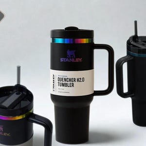 May include: Three black Stanley Quencher H2.0 tumblers with handles and straws. The central tumbler features a white product label reading Stanley Quencher H2.0 Tumbler. Each tumbler has a metallic iridescent ring below the black lid.