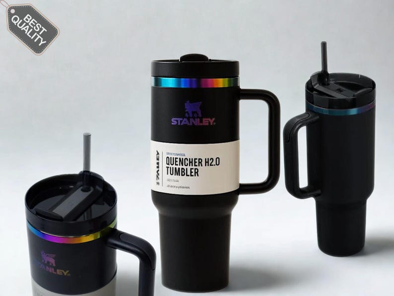 May include: Three black Stanley Quencher H2.0 tumblers with handles and straws. The central tumbler features a white product label reading Stanley Quencher H2.0 Tumbler. Each tumbler has a metallic iridescent ring below the black lid.
