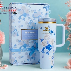 May include: A light blue Stanley tumbler and matching gift box, both adorned with a floral pattern in white and blue. The tumbler has a handle and a gold rim. The box has a blue ribbon and the text "US STOCK FAST DELIVERY Ship from USA".